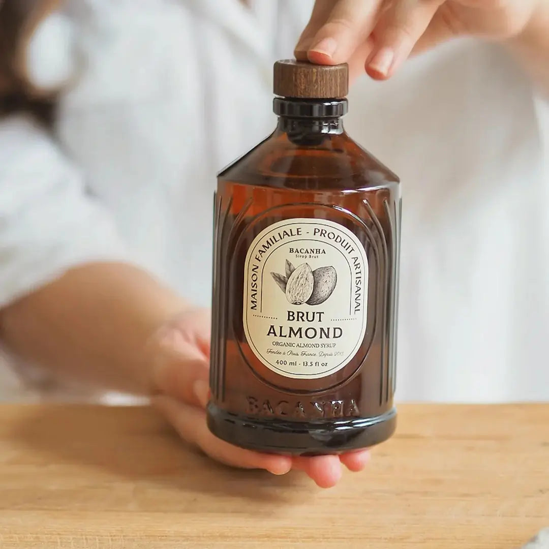 Szklana butelka organicznego syropu migdałowego Bacanha Brut Almond na drewnianym blacie z migdałami w tle.