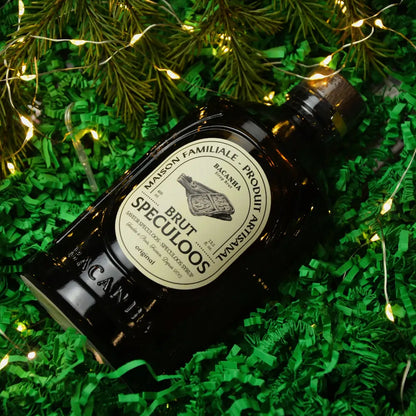 Butelka syropu Bacanha Brut Speculoos leżąca na zielonym wypełnieniu, otoczona gałązkami świerku i świątecznymi światełkami.