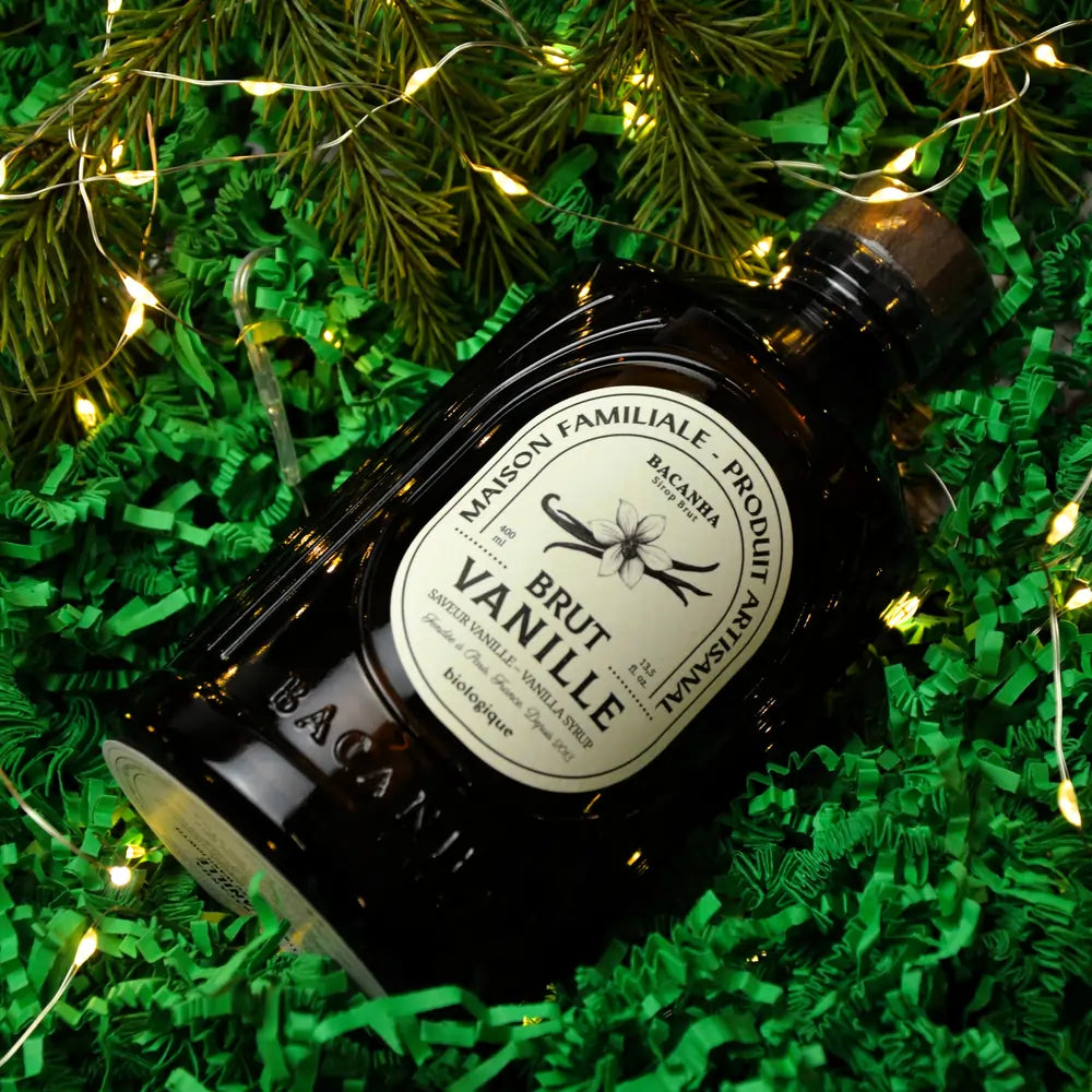 Butelka syropu Bacanha Brut Vanille ułożona na zielonym wypełnieniu, otoczona gałązkami świerku i świątecznymi światełkami.