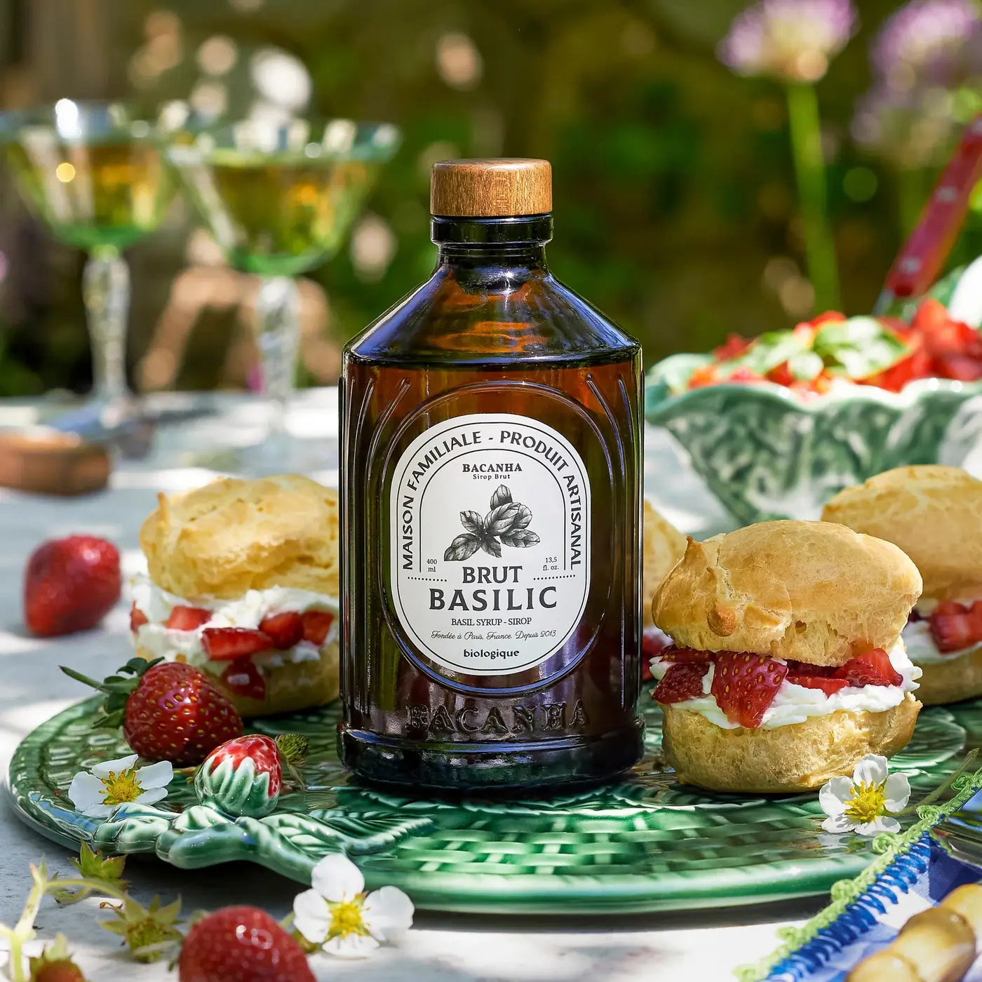 Naturalny syrop bazyliowy BACANHA w butelce, podany na zielonym talerzu z truskawkami i bułeczkami, idealny do napojów i deserów.
