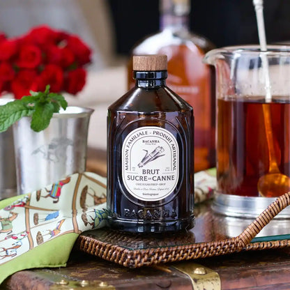 Butelka syropu z trzciny cukrowej o nazwie Brut Sucre de Canne ustawiona na stoliku obok innych akcesoriów barmańskich i czerwonych kwiatów