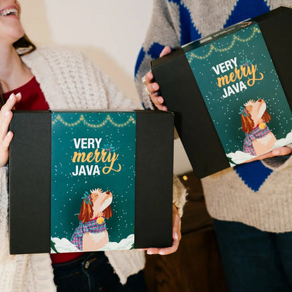 Świąteczne pudełko prezentowe Very Merry JAVA w czarnym opakowaniu z zieloną kartonową owijką, trzymane przez dwie osoby w zimowych swetrach.