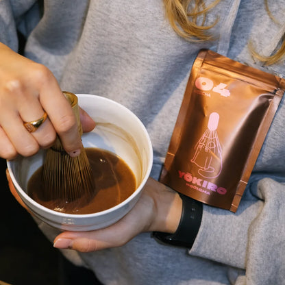 Lifestyle zdjęcie przygotowania hojichy Yōkiro – ubijanie hojichy bambusową miotełką w czarce, opakowanie hojichy w tle