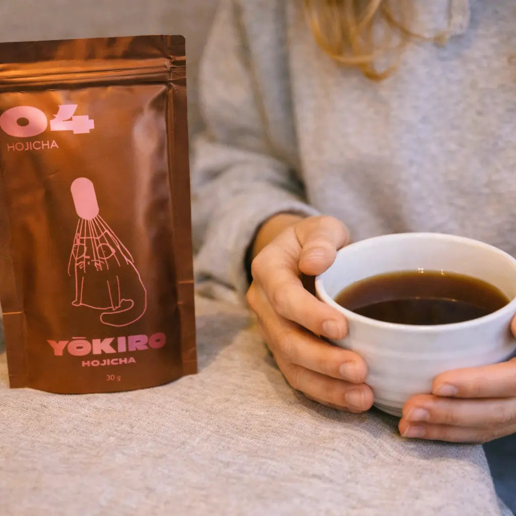 Lifestyle zdjęcie hojichy Yōkiro – opakowanie hojichy obok białej czarki z zaparzoną hojichą trzymanej w dłoniach