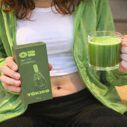 Lifestyle zdjęcie matchy Yōkiro Ceremonial – kartonowe opakowanie 30 g trzymane w dłoni oraz zielona matcha w szklance