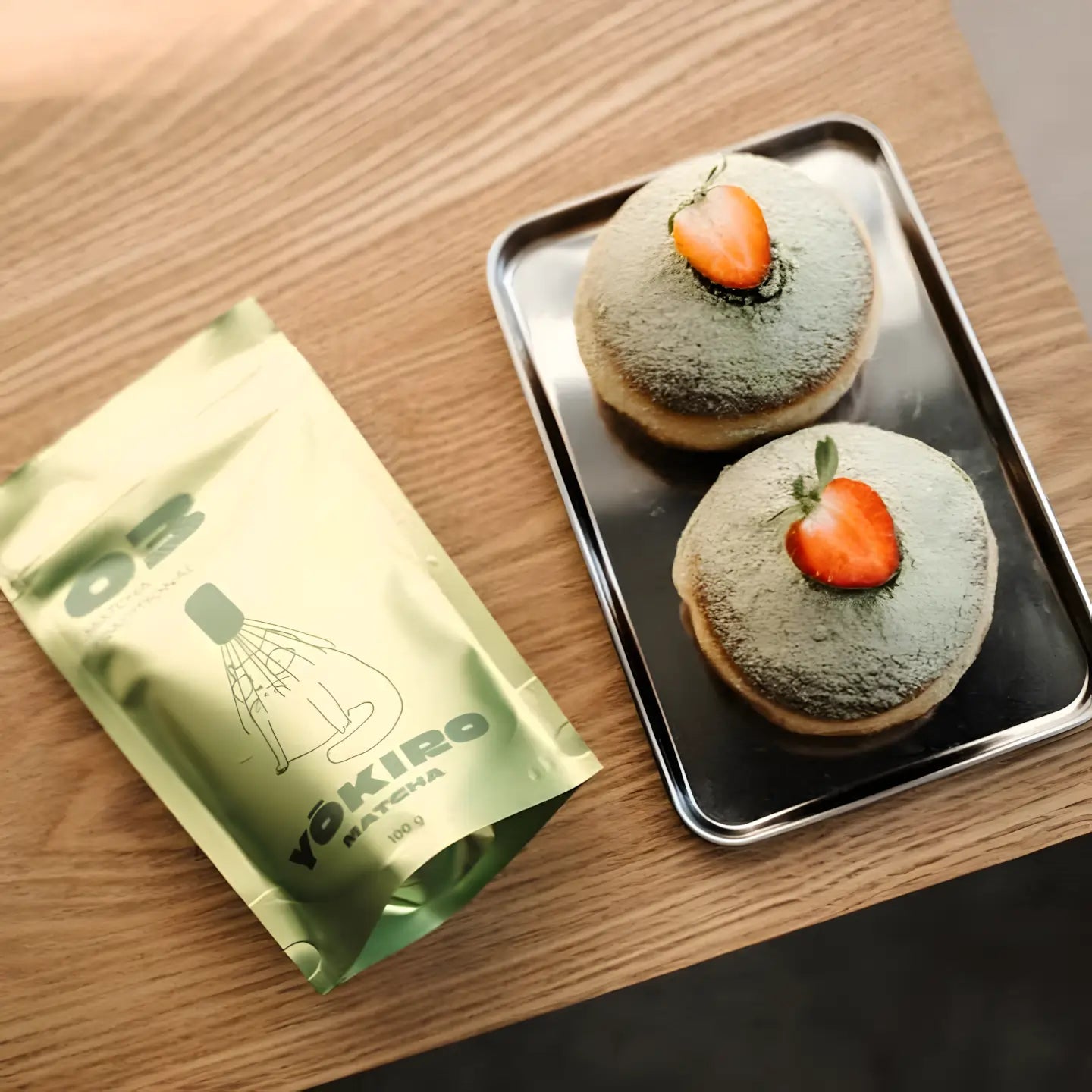 Lifestyle flat lay z matchą Yōkiro Traditional – opakowanie matchy obok pączków z matchą i truskawką na drewnianym stole