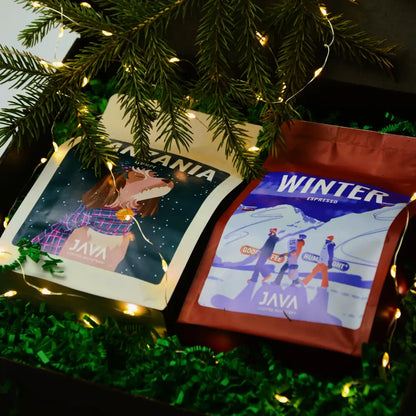 Dwie kawy JAVA Coffee – Tanzania Itumpi i Winter Espresso – ułożone w świątecznym pudełku na zielonym wypełnieniu, ozdobione światełkami i gałązkami świerku.