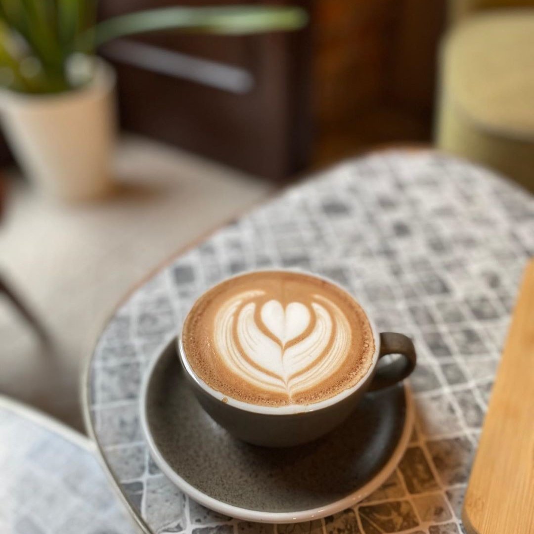 Filiżanka cappuccino z latte art w kształcie serca, stojąca na stoliku w kawiarni o przytulnym, ciepłym wnętrzu.